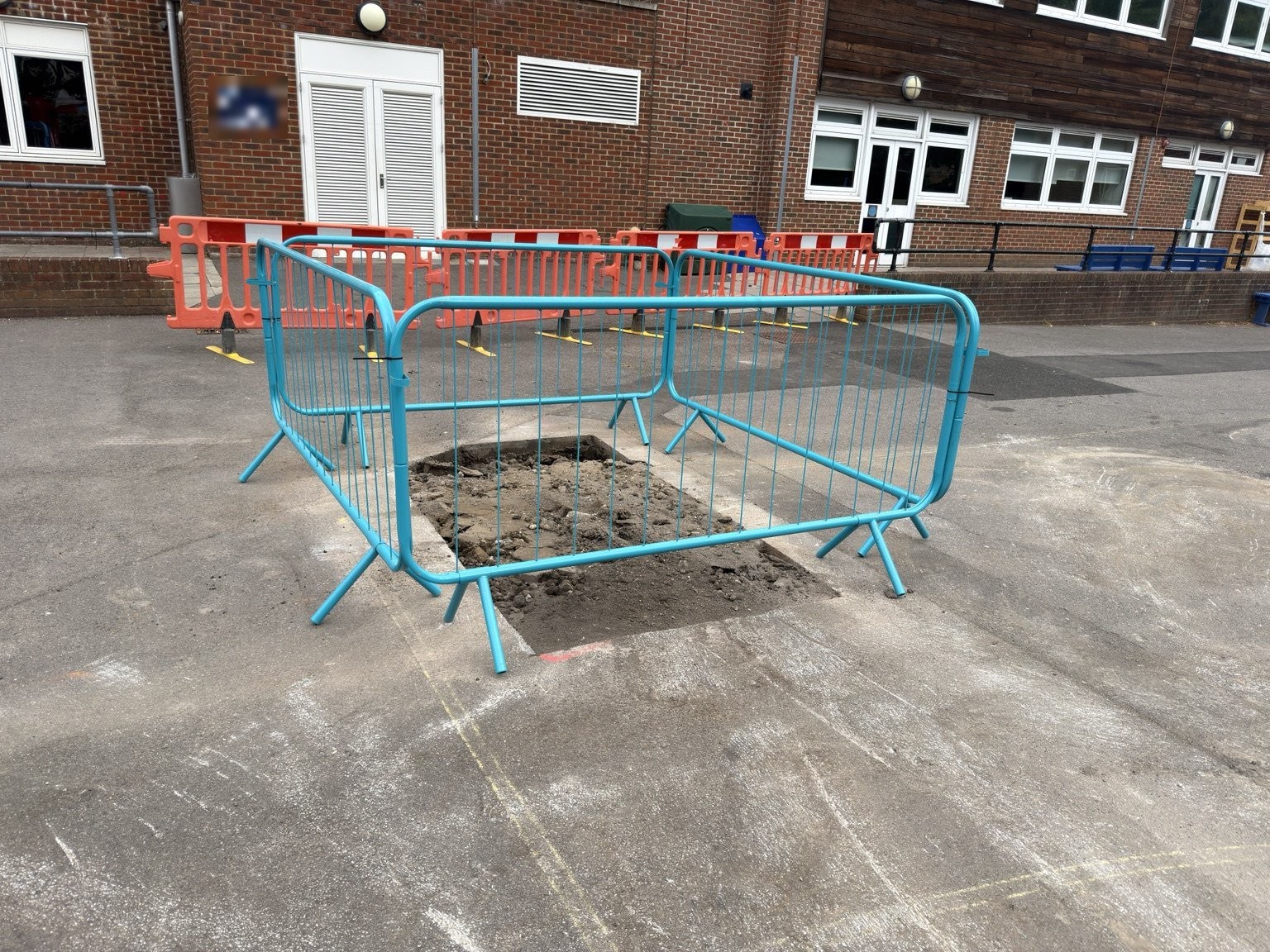 Excavated trial pit in a hard-surfaced area surrounded by blue safety barriers, with orange barriers and a brick building behind